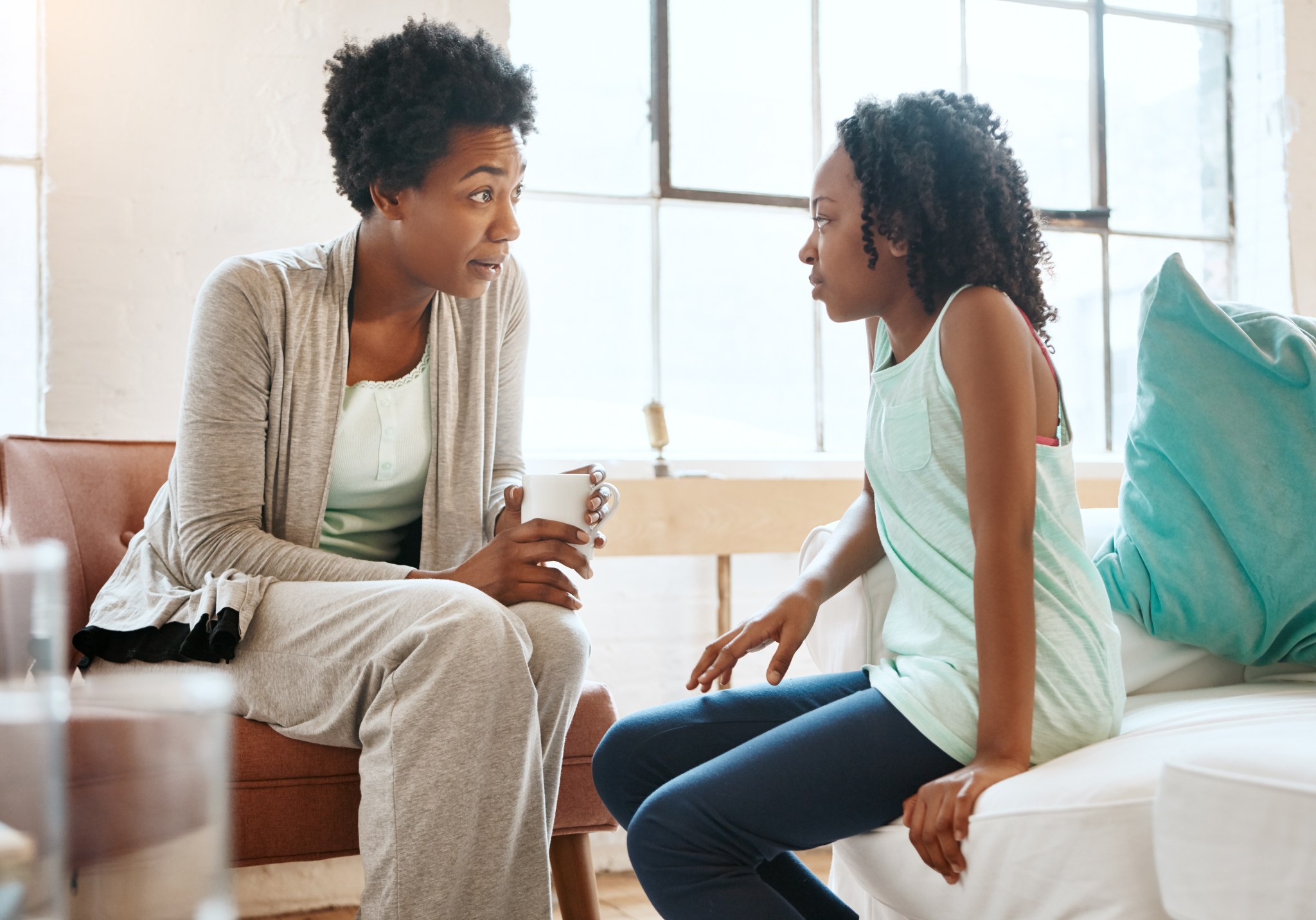 Une femme et une jeune fille assises dans un salon lumineux, discutant ensemble. La femme tient une tasse, tandis que la jeune fille l'écoute attentivement. Cette image évoque des moments de soutien et de partage, en lien avec le thème du cancer du sein.