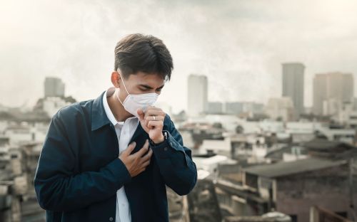 Photographie d’un homme portant un masque respiratoire, en train de tousser au milieu d’une pollution urbaine dense.